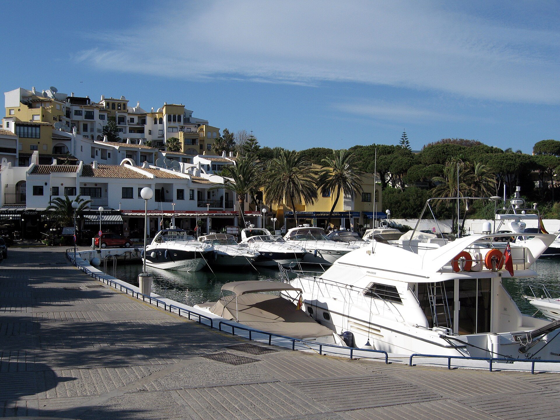 The harbour village at Cabopino, Marbella, boats moored along the quay with stepped white buildings around the water. Photograph: Olaf Tausch, CC BY 3.0.