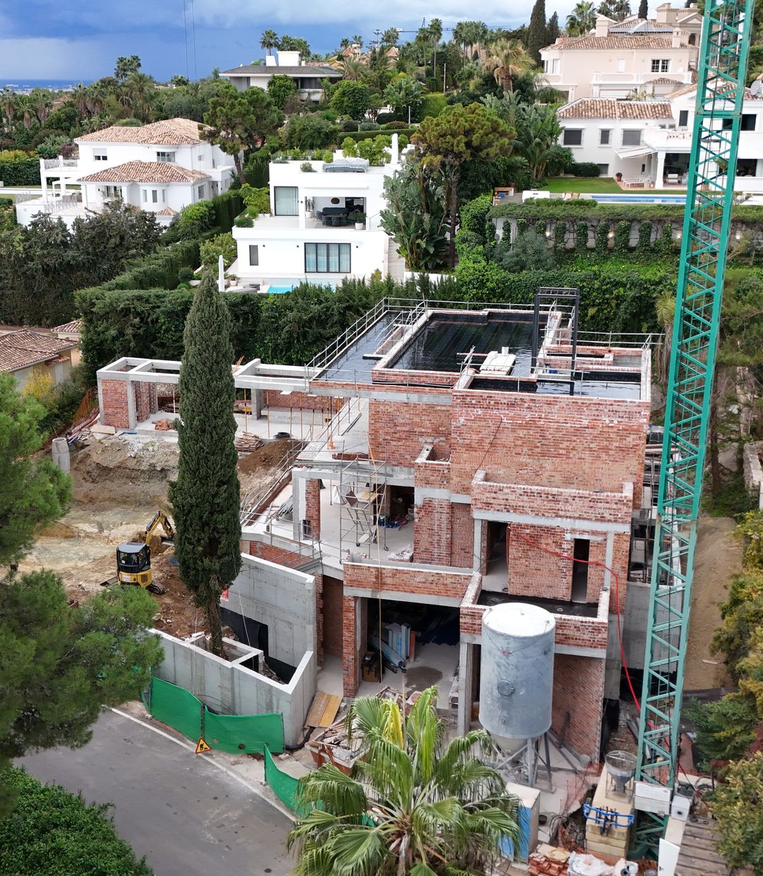 Aerial view of a Marbella villa under construction, its brick and concrete shell open to the sky beside a tower crane, surrounded by completed white villas and mature gardens
