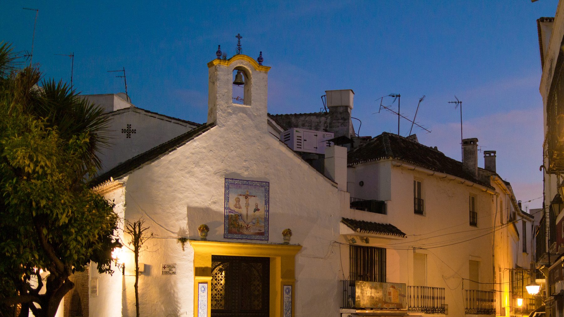 The whitewashed Santo Cristo chapel and its bell tower at dusk in Marbella Old Town, tiled roofs stepping away behind it